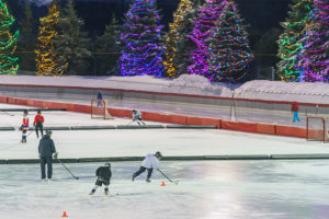 Photo of children playing ice hockey with trees lit with holiday lights in the background